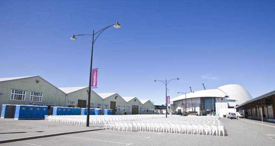Rows of white chairs and blue portable toilets lined up in a large concrete area with the white curved roof of the Maritime Museum in the background.