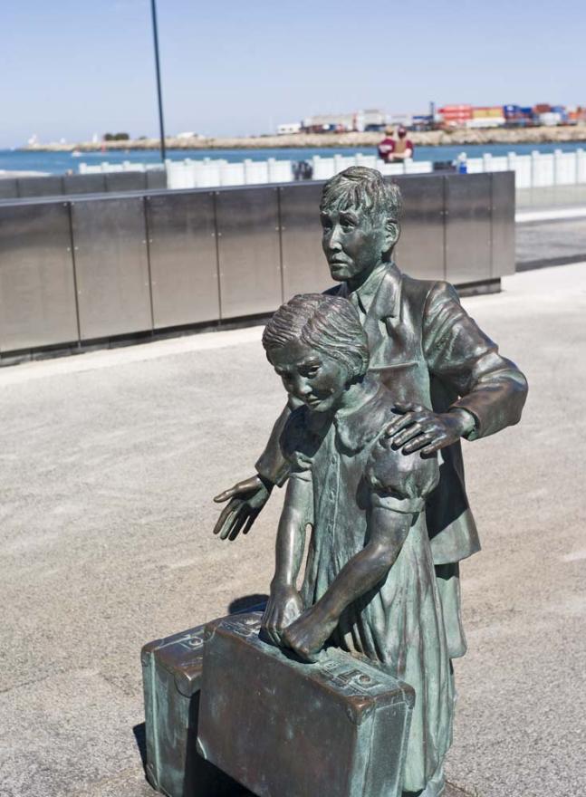 A statue of a young boy and girl holding a suitcase with the ocean visible in the background. The statue appears green where it was corroded.