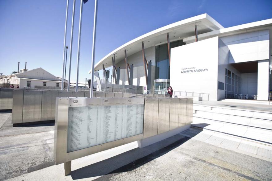 Exterior of the Maritime Museum with glass entrance and flagpoles in front. A series of metal walls engraved with names are visible in the foreground