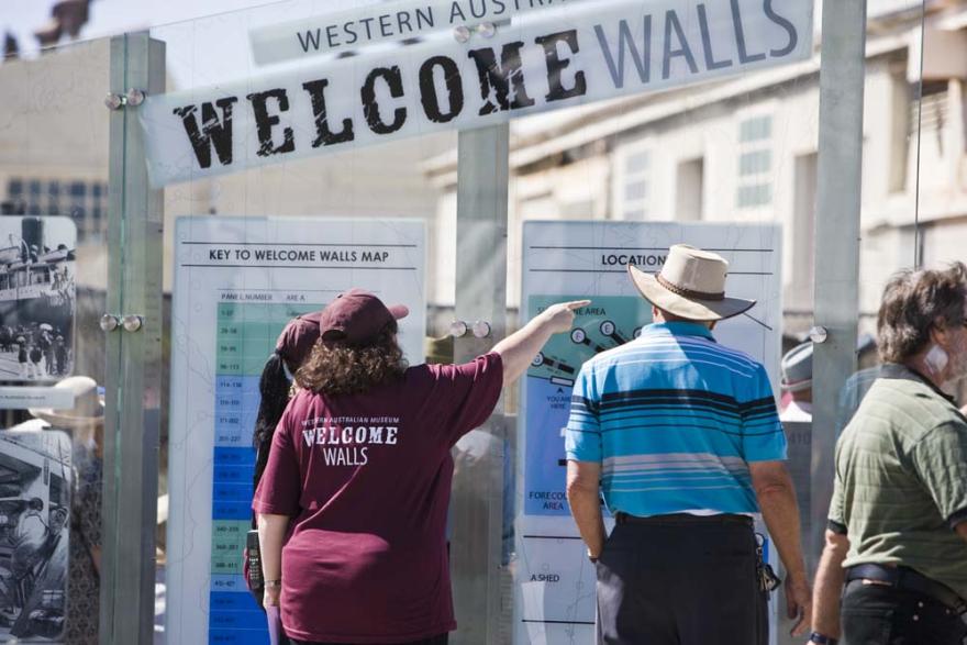 A staff member in a dark red uniform with the words Western Australian Museum Welcome Walls points to something off camera while assisting individuals in front of a glass map display