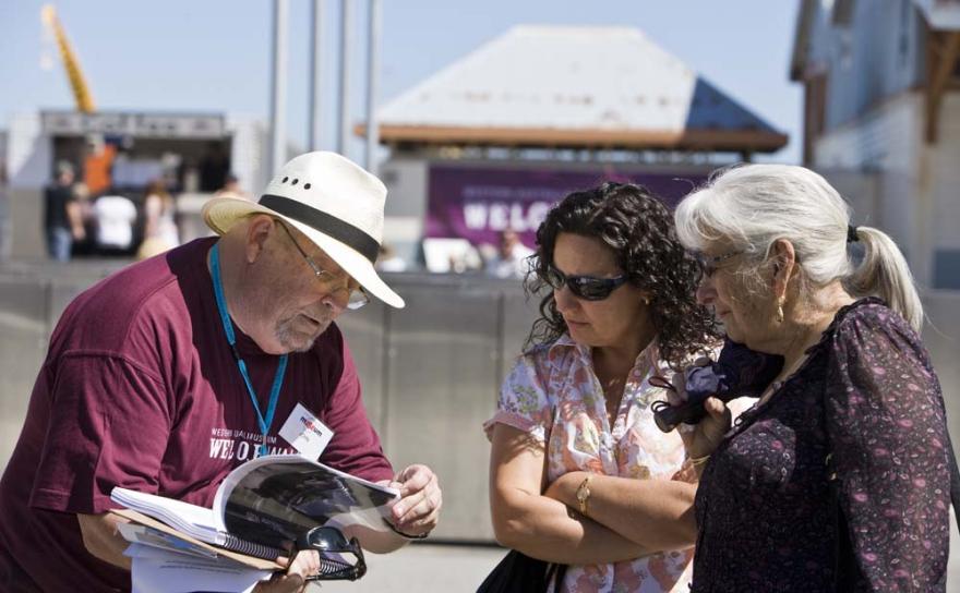 A staff member in a dark red uniform with the words Western Australian Museum Welcome Walls holds open a book while two individuals look on