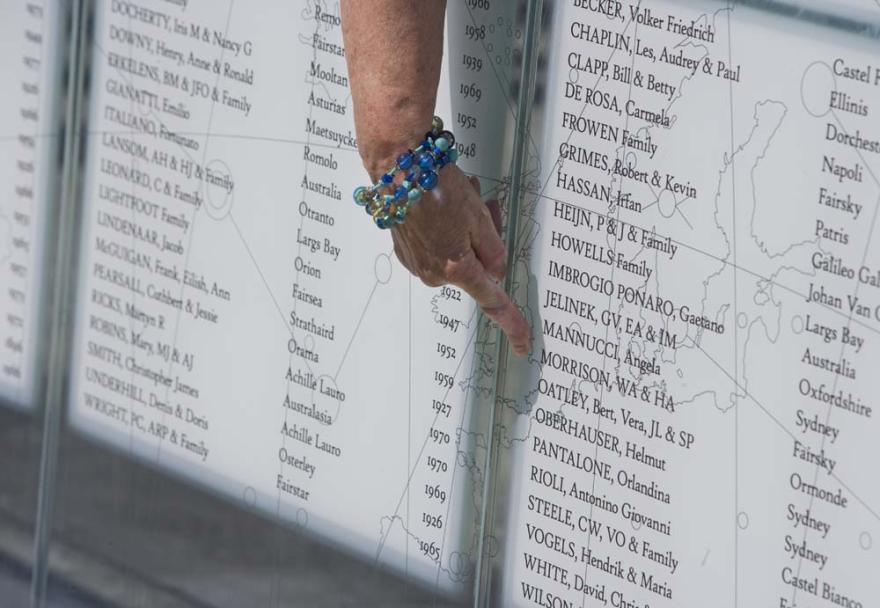 A hand wearing a bracelet of blue beads points to a name on an engraved glass panel
