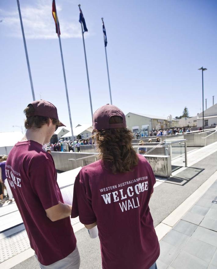 Two staff members in dark red uniform shirts with the words Western Australian Museum Welcome Walls face away from the camera and towards a large concrete area with flagpoles on a sunny day