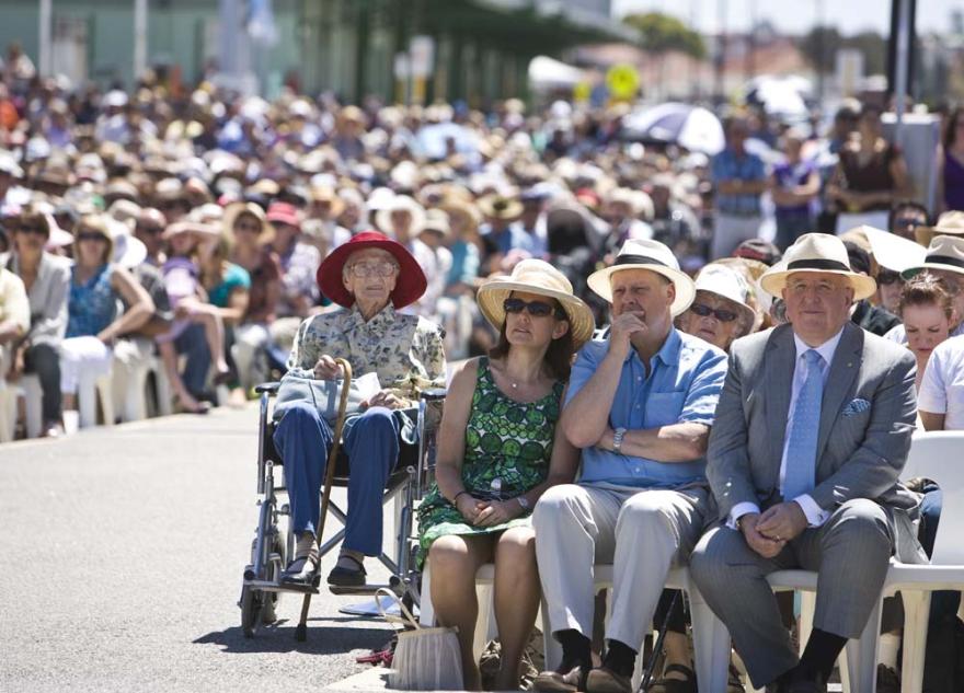 Rowds of individuals seated on white plastic chairs stretch back across a wide open concrete space