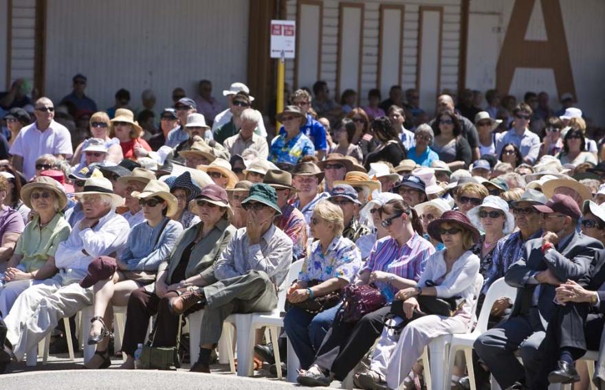 Rowds of individuals seated on white plastic chairs stretch back across a wide open concrete space