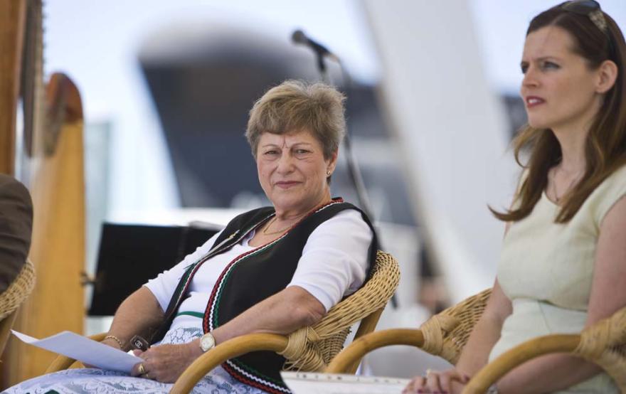 An individual wearing cultural dress sits in a wicker chair holding paper while another person in a green shirt is visible next to them and out of focus