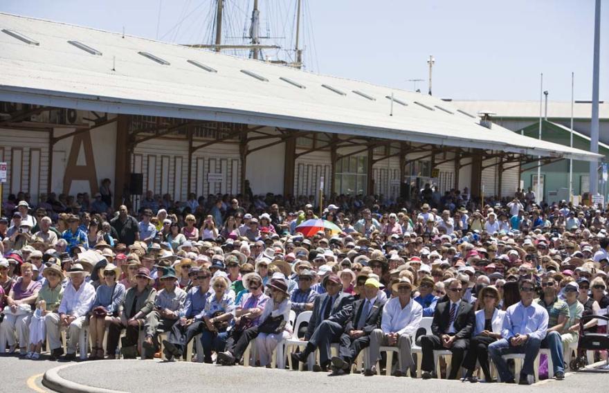 A crowd of people sat in rows on plastic white chairs in a large concreted space next to a big maritime shed with an 'A' on it.