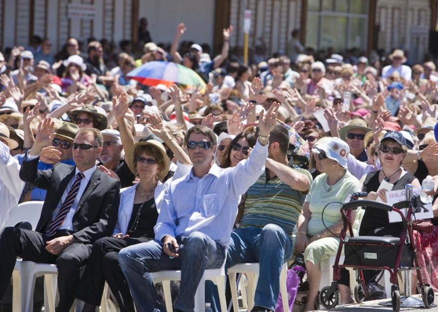 A crowd of people sat in rows on plastic white chairs in a large concreted space and hold their hands up as though waving