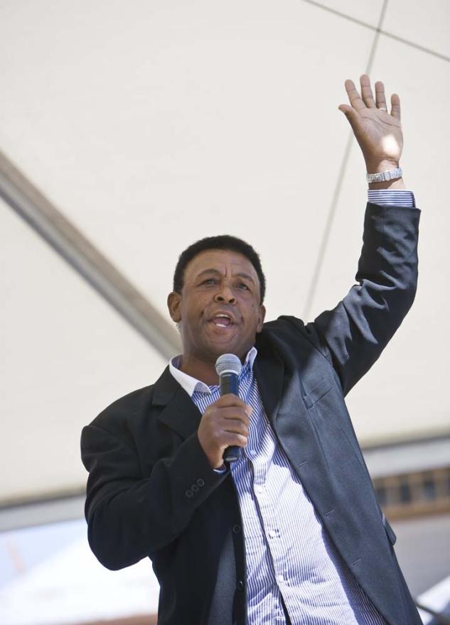 An individual in a black suit holds a microphone in one hand and raises the other as though waving while talking with an animated expression