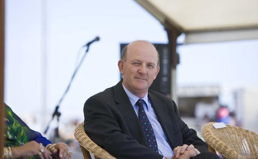 A person wearing a black pinstripe suit sits in a wicker chair and watches something off camera intently