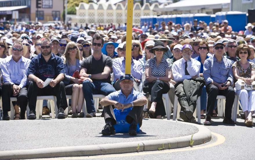 An individual sits on a kerb leaning against a yellow signpost while a crowd of people sat in rows on plastic white chairs in a large concreted space is visible behind them