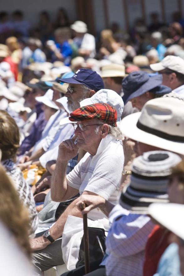 An individual wearing a red tartan hat is pictured sitting among a crowd of people sat in rows on plastic white chairs in a large concreted space
