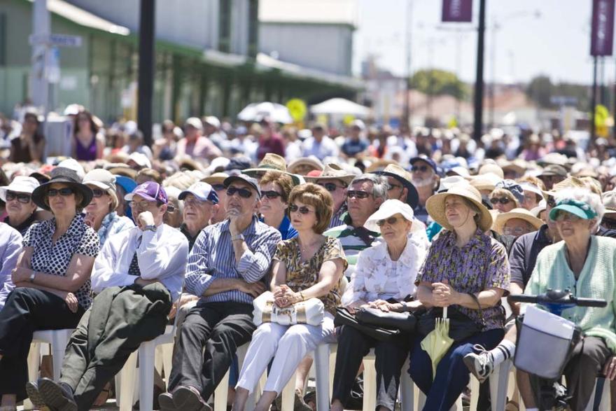 A crowd of people sat in rows on plastic white chairs in a large concreted space