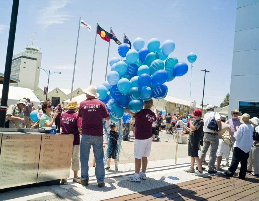 Several individuals in dark red shirts that read Western Australian Museum Welcome Walls hold a large bunch of vibrant blue balloons in front of a busy event area with white buildings in the background