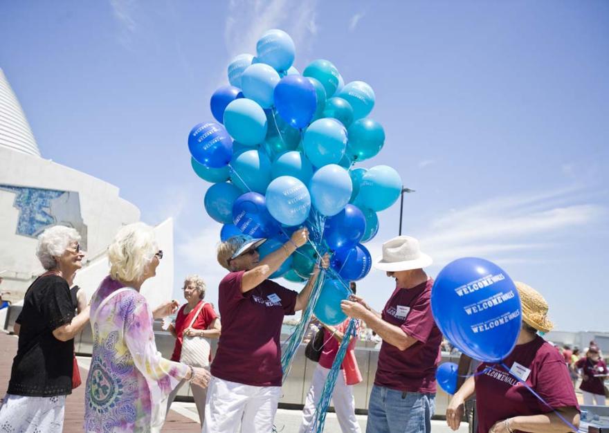 Several individuals in dark red shirts that read Western Australian Museum Welcome Walls hold a large bunch of vibrant blue balloons in front of the Western Australian Maritime Museum
