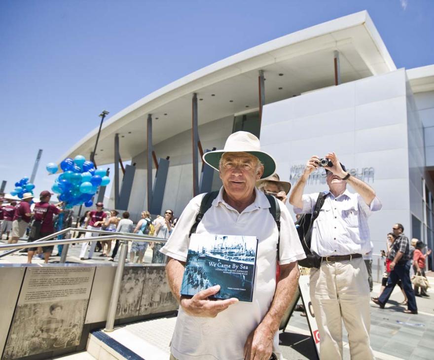 A person in a white polo shirt and wide brimmed hat stands in front of the WA Maritime Museum and holds a book entitled We Came By Sea for the camera