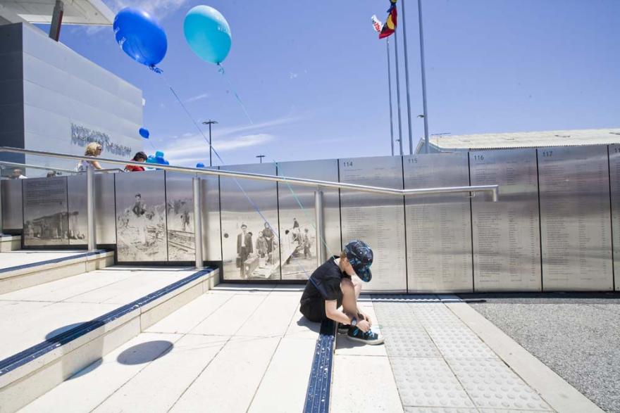 A young child sits on a step to tie their shoelaces while holding two blue balloons