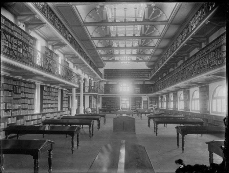 A grand, historic library interior with tall bookshelves, a second-floor balcony, and large wooden tables.