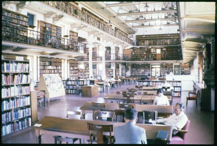 Interior of a large library with two levels of bookshelves, wooden tables, and natural lighting.