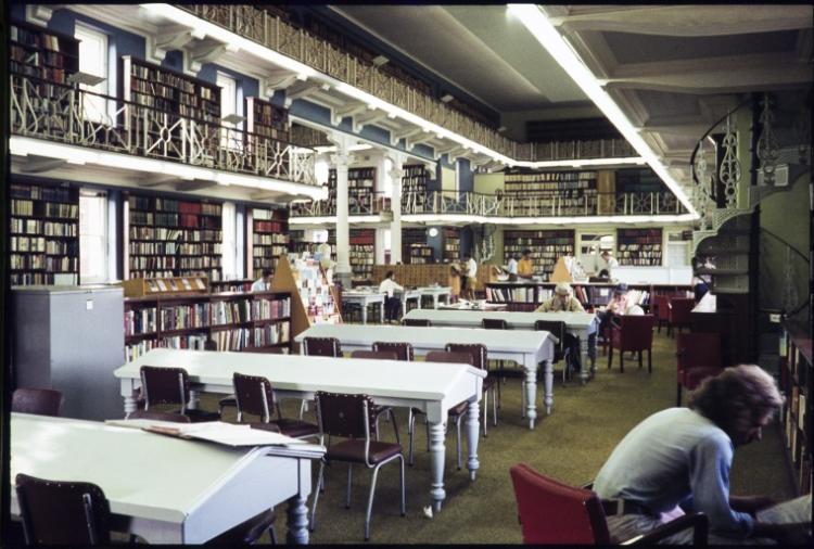 Interior of a two-level library with bookshelves, white tables, and people reading.