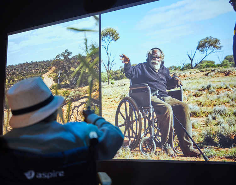 A person in a wheelchair looks up at a screen projecting a film set in Spinifex Country