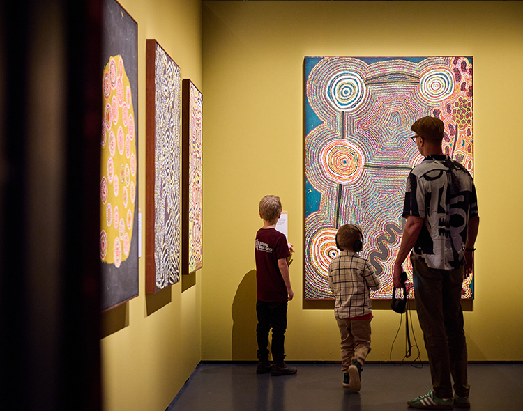 An adult and 2 children inspecting a Spinifex artwork on display in Gallery at WA Museum Boola Bardip