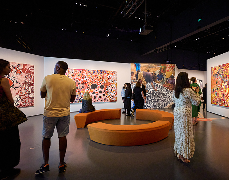 People stand in an exhibition space with circular seating modules in the centre of the room, surrounded by artworks and images from Spinifex country