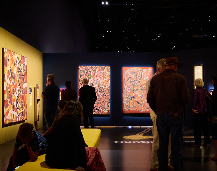 People observing Spinifex artworks in a darkly lit Gallery space