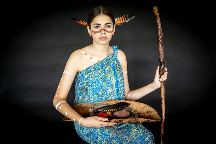 A young person wearing a blue and yellow dress with brown and yellow body paint holding a long sculptured staff and a wooden bowl of cultural materials 