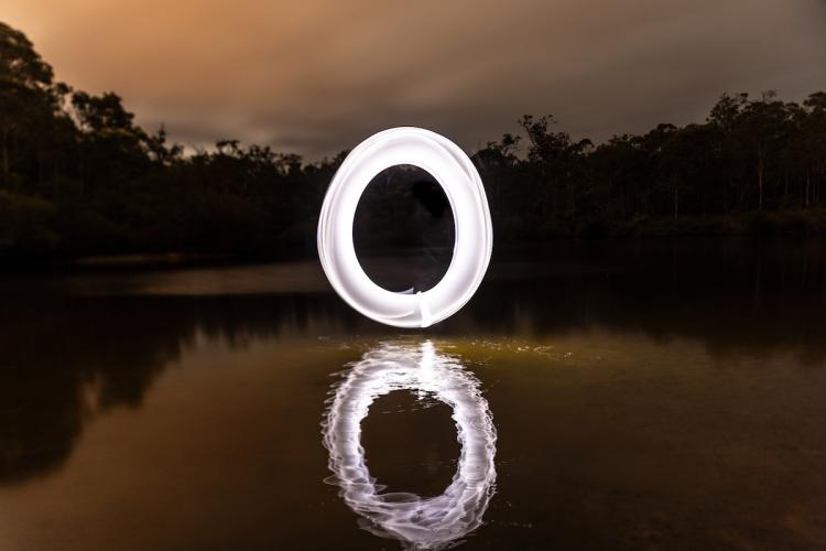 A circular white light sculpture hovering above a lake on a dusk evening