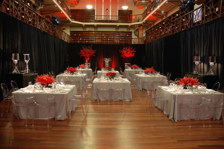 Elegant dining setup with white tablecloths, red flower centerpieces, and transparent chairs in a large room with black drapes.