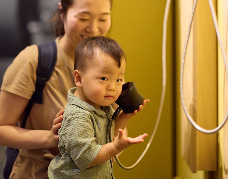 A young child holds an audio puck to his ear with an adult smiling behind him.