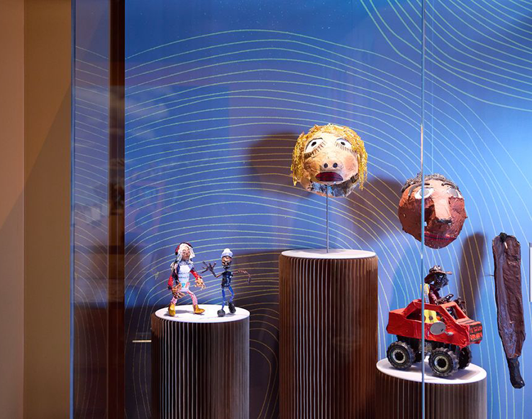 Museum exhibition display with colourful figures, large heads, and a car on pedestals.