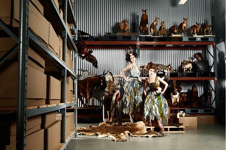 Two models in feathered dresses stand in a storage room with taxidermied animals on shelves.