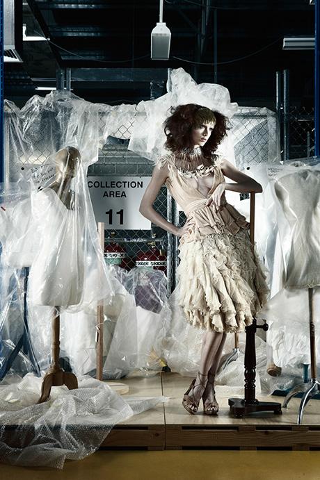 Model in a ruffled cream dress posing in a storage area with plastic-covered dress forms.