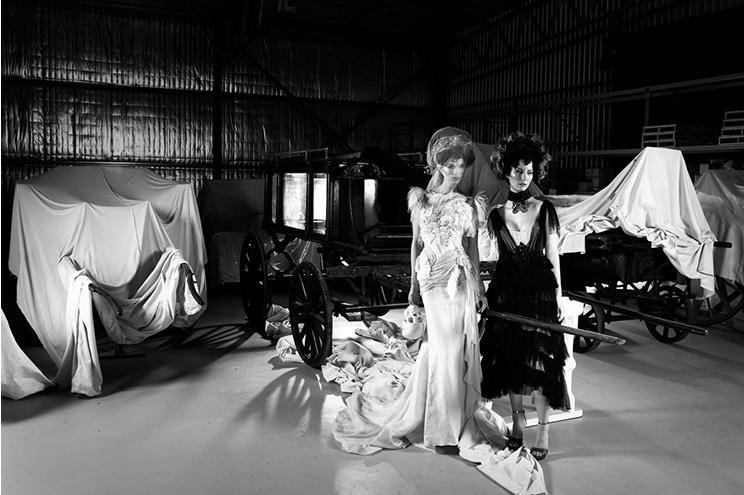 Two models in elaborate gowns stand in front of partially covered carriages in a dimly lit storage space.