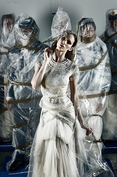 A fashion model in an embellished gown stands in front of figures wrapped in bubble wrap.