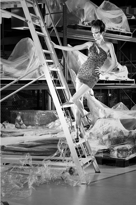 Model posing on a ladder in a warehouse with items covered in plastic sheets.
