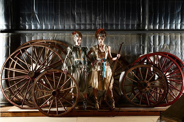 Two people in vintage-style clothing stand among large wooden wagon wheels against a metallic wall.