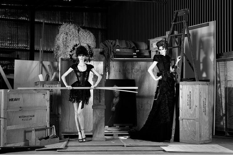 Two models in elaborate black outfits pose in a warehouse among wooden crates and ladders.