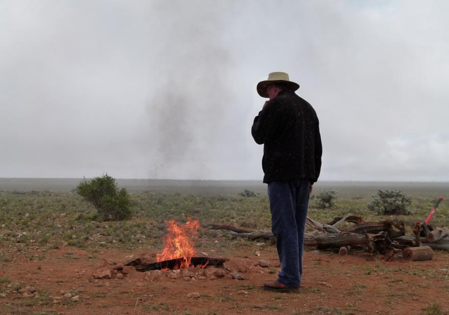 A person wearing a hat stands beside a campfire, logs and an axe visible in the background, in an expansive plains environment
