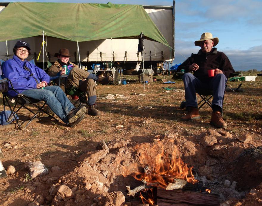 3 people sit in chairs in front of a campfire, their campsite in the background