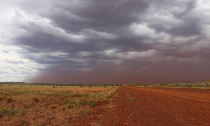 Red dirt tinged storm clouds roll in across a grassland landscape, a red dirt road rolling through the centre of the image.