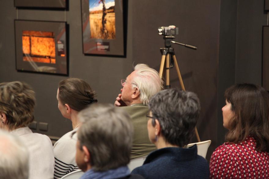 An audience listening in a room with framed artworks on the wall and a camera on a tripod.