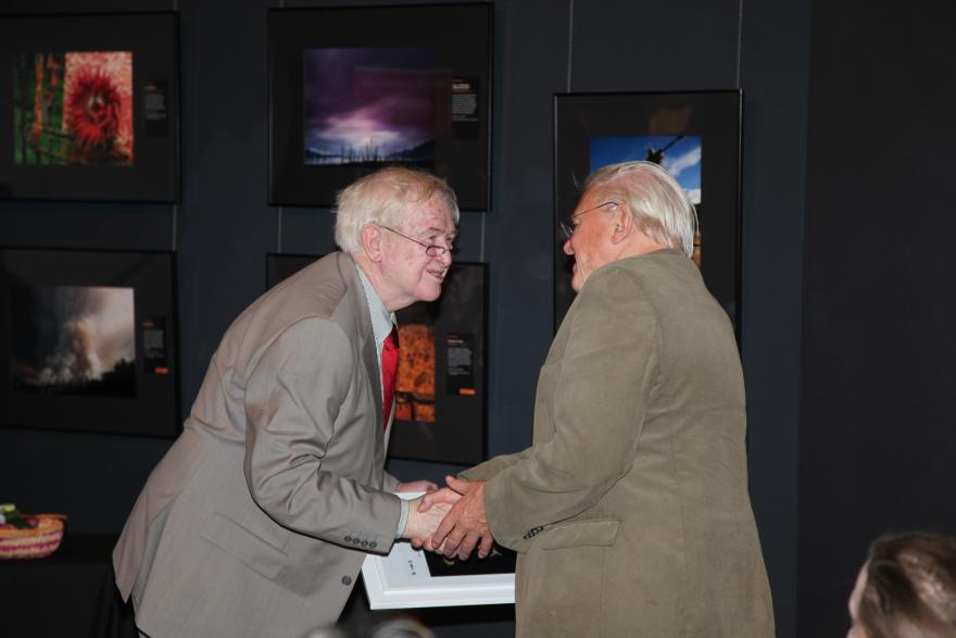Two people wearing suit jackets shake hands in a dimly lit room, as one presents the other with a framed picture.