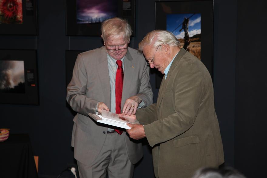 Two people wearing suit jackets stand side by side in a dimly lit room, as they examine a framed picture.