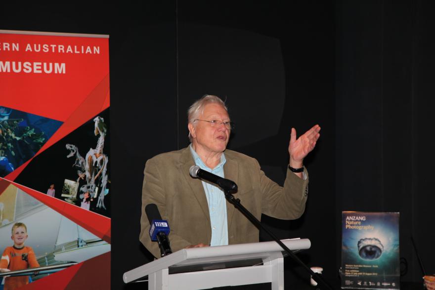 Person speaking at a podium with a red banner and a nature photography poster in the background.