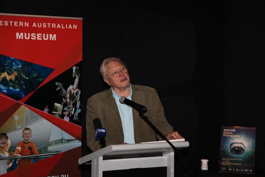 Person speaking at a podium with a red banner and a nature photography poster in the background.