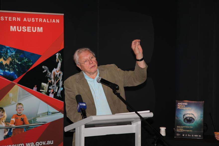 Person speaking at a podium with a red banner and a nature photography poster in the background.