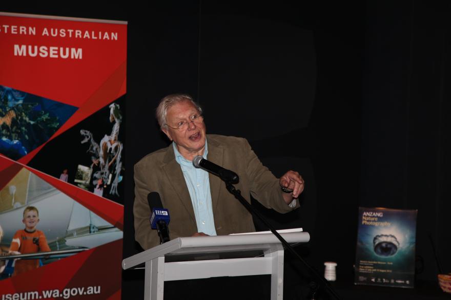 Person speaking at a podium with a red banner and a nature photography poster in the background.